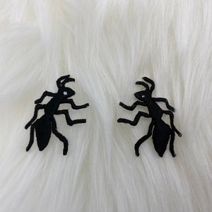 May include: Two black embroidered ant appliques on a white fuzzy background.