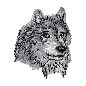 May include: An embroidered patch featuring the head of a wolf in shades of silver, gray, and white. The design includes detailed black stitching to define the fur texture, pointed ears, alert eyes, and a dark nose on a white background.