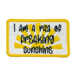 May include: A rectangular patch with a white background and a yellow border. The patch features the text "I AM A RAY OF FREAKING SUNSHINE" in black, with a yellow sunburst design.
