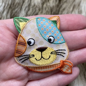 May include: A colorful embroidered iron-on patch featuring a cartoon cat with a yellow nose, orange and white stripes, and a blue and white polka dot scarf.