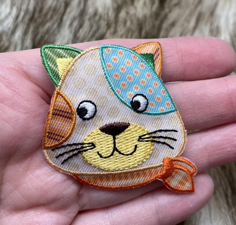 May include: A colorful embroidered iron-on patch featuring a cartoon cat with a yellow nose, orange and white stripes, and a blue and white polka dot scarf.