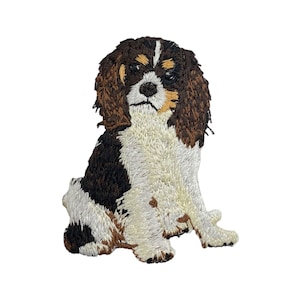 May include: Embroidered patch of a brown and white Cavalier King Charles Spaniel sitting with its head tilted to the side.