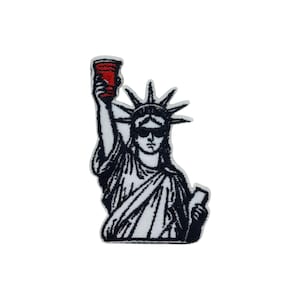 May include: Black and white embroidered patch of the Statue of Liberty wearing sunglasses and holding a red plastic cup.