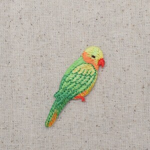 May include: A green and yellow embroidered bird applique with a red beak and orange feet. The bird is on a beige fabric background.