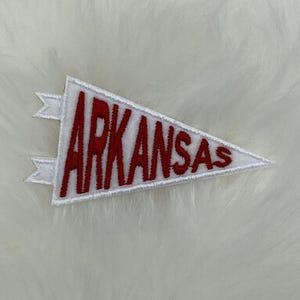 Peut inclure: Un fanion en feutre blanc avec du texte brodé en rouge qui dit "ARKANSAS".