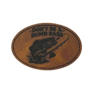 May include: Oval-shaped, brown leather patch with the text "DON'T BE A DUMB BASS" above a black graphic of a fish jumping out of the water with a fishing rod. The patch has a stitched border.