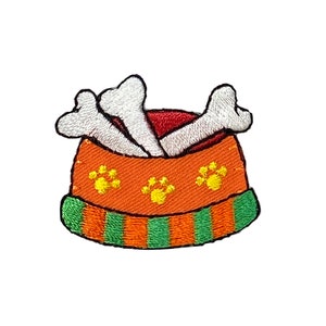 May include: An embroidered patch of a dog bowl with two white bones inside. The bowl is orange with green and orange stripes around the rim and has three yellow paw prints on the front.