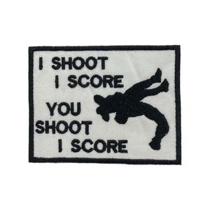 May include: A rectangular white patch with black text that reads "I SHOOT I SCORE YOU SHOOT I SCORE". A black silhouette of a person is shown in the lower right corner. The patch has a black border.