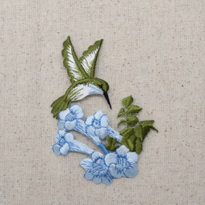 Puede incluir: Parche bordado con un colibrí con alas verdes y blancas, un pico negro y un cuerpo verde. El colibrí está posicionado sobre flores azul claro con costuras detalladas y hojas verdes. El parche está sobre un fondo de tela beige.