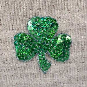 May include: A green shamrock-shaped patch made of sequins. The sequins are arranged in a pattern that resembles a four-leaf clover.