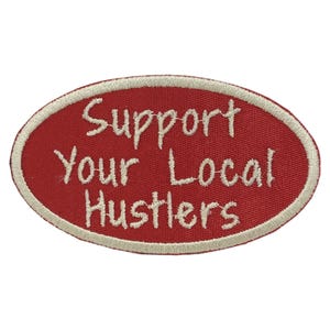 May include: Oval-shaped red patch with white embroidered text that reads "Support Your Local Hustlers." The patch has a white border and is likely designed to be sewn or ironed onto clothing or accessories.