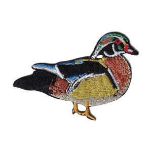 May include: Embroidered patch of a colorful wood duck. The patch features detailed stitching in shades of green, blue, red, yellow, and brown. The duck is depicted in a side profile, with its head turned slightly.