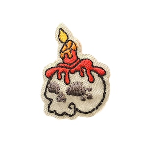 Puede incluir: Parche bordado de una calavera blanca con una vela roja en la parte superior. La vela tiene una llama amarilla y cera roja goteando por la calavera.