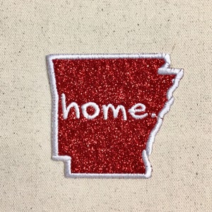 May include: Red glitter iron-on patch shaped like the state of Arkansas with white stitching around the border and the word "home" in white lettering.