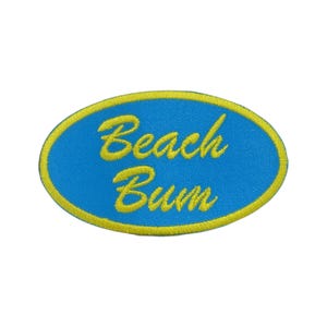 May include: An oval-shaped embroidered patch featuring a bright blue background with a yellow border. The text Beach Bum is stitched in yellow cursive lettering across the center of the patch, creating a clear contrast against the blue fabric.