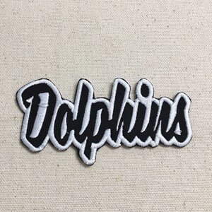 May include: A black and white embroidered patch with the word "Dolphins" in a stylised font.