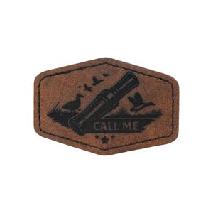 Puede incluir: Un parche de cuero marrón con un gráfico negro de un reclamo de pato, patos y el texto "CALL ME". El parche tiene forma hexagonal con un borde cosido y detalles de estrellas.