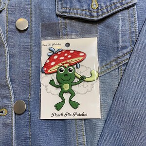 Puede incluir: Parche para planchar con una rana verde que sostiene un paraguas de hongo rojo y blanco. El parche está en una tarjeta blanca con el texto "Iron On Patches" y "Peach Pie Patches".