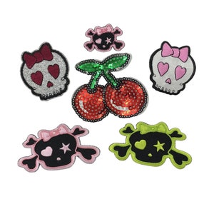 Puede incluir: Cinco parches para planchar con diseños de calavera y huesos cruzados. Dos parches son rosa y blanco con purpurina y un lazo. Un parche es negro y verde con una estrella y un lazo. Dos parches son cerezas rojas y verdes con lentejuelas.