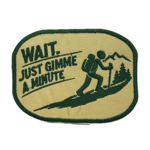 May include: Embroidered patch with a beige background and a green border. The patch features the text "WAIT. JUST GIMME A MINUTE" above a graphic of a hiker with a backpack, trekking pole, and trees. The hiker is ascending a hill.
