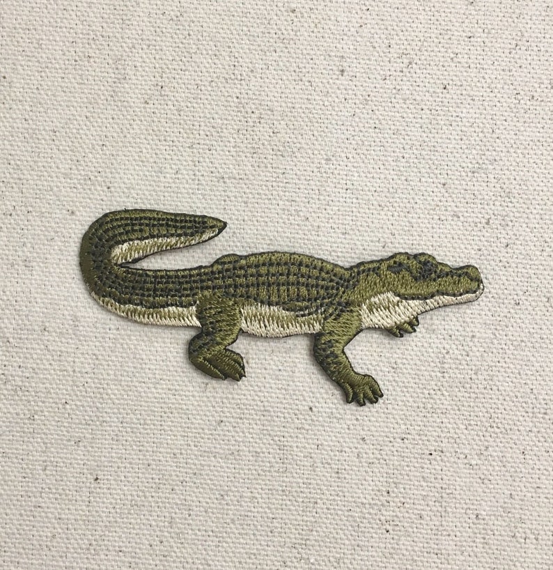 May include: Green embroidered alligator patch. The alligator is facing right and has a textured, scaly appearance.