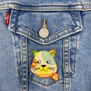 May include: A colorful patchwork cat applique with a yellow face, orange nose, and blue eyes is sewn onto a blue denim jacket pocket.