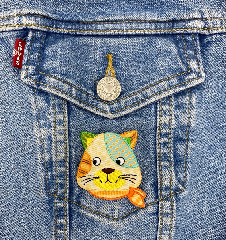 May include: A colorful patchwork cat applique with a yellow face, orange nose, and blue eyes is sewn onto a blue denim jacket pocket.