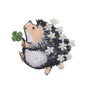 Könnte beinhalten: Gestickter Bügelaufkleber mit einem niedlichen Igel, der einen Kleeblatt in seiner Pfote hält und weiße Blüten auf seinem Rücken hat.