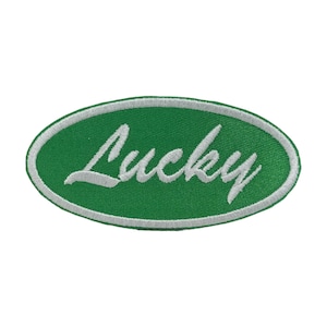 May include: Green oval patch with the word "Lucky" embroidered in white script. The patch has a white border and is set against a white background. The patch is likely for clothing or accessories.