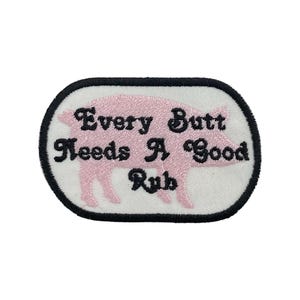 May include: Embroidered patch with a pink pig silhouette and the text "Every Butt Needs A Good Rub" in black script. The patch has a white background with a black border, suitable for clothing or accessories.