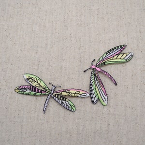 May include: Two embroidered dragonfly patches with colorful wings in shades of pink, green, yellow, and white. The dragonflies have detailed antennae and bodies, set against a neutral beige fabric background.