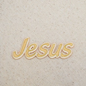 Könnte beinhalten: Ein beigefarbener Stoffhintergrund mit dem Wort "Jesus" in kursiv geschriebener Schrift gestickt. Die Buchstaben sind mit goldfarbenem Garn umrandet und mit einer hellbeigen Farbe gefüllt.
