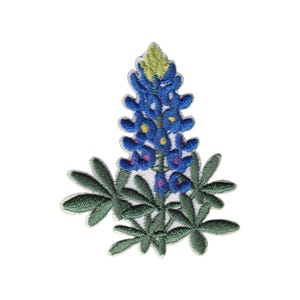 May include: An embroidered patch featuring a bluebonnet flower with vibrant blue petals, a pale green tip, and dark green palmate leaves. The design is rendered in detailed thread work with small yellow and pink accents on the flower blossoms.