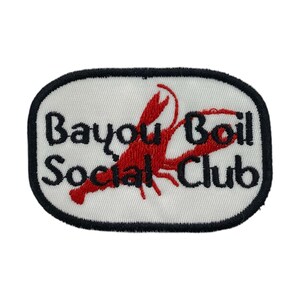 Op de afbeelding: Geborduurde patch met de tekst "Bayou Boil Social Club" in zwart, met een rood rivierkreeftontwerp op een witte achtergrond. De patch heeft een zwarte rand en een ovale vorm.