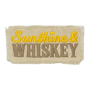May include: A beige fabric patch with frayed edges, featuring the embroidered text "Sunshine & Whiskey" in yellow and brown thread.