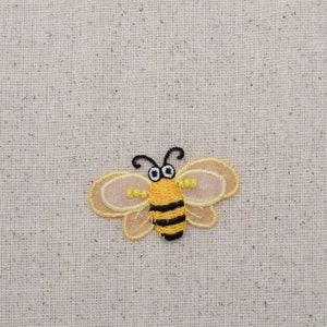 May include: A yellow and black embroidered bee with translucent wings. The bee is on a beige fabric background.