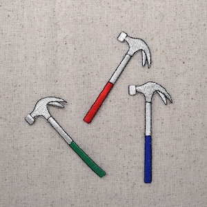May include: Three embroidered hammer patches. Each hammer has a silver head and a handle with a different color: red, green, and blue. The patches are on a beige fabric background.