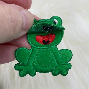 Puede incluir: Aplique de rana de fieltro verde con boca roja y ojos blancos. La rana está cosida con hilo negro y tiene una sonrisa cosida.