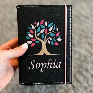 Könnte beinhalten: Schwarzer Filz-Reisepasshalter mit einem Baumdesign, bestickt in Rosa, Türkis und Weiß. Der Name "Sophia" ist darunter in weißer Schrift gestickt. Ein rosa Gummiband sichert den Halter.