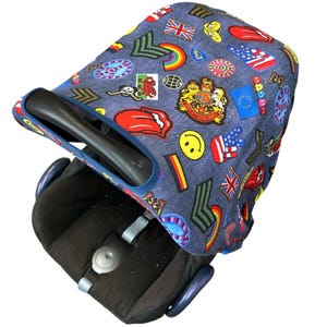 May include: A denim-effect baby car seat cover featuring a variety of colourful patches, including flags, a rainbow, and a smiley face. The cover is over a black car seat with a black handle and blue accents.