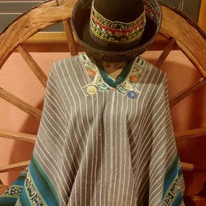 May include: A gray and white striped poncho with blue and green accents and embroidered details. A black hat with colorful trim sits atop the poncho. The poncho is displayed on a wooden wagon wheel.