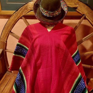 May include: A vibrant red poncho with patterned blue, green, and white accents on the sleeves. A brown hat with a patterned band sits atop the poncho. The poncho is displayed on a mannequin, showcasing its design and texture.