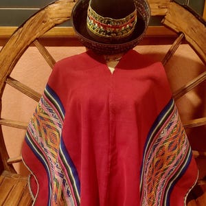 May include: A red poncho with colorful geometric patterns on the sleeves and a matching hat. The poncho has a V-neck and is displayed on a wooden stand. The hat is dark brown with a decorative band.