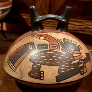 May include: A rounded ceramic vessel with a bridge handle and two spouts. The surface is painted in shades of tan, black, and terracotta, featuring a stylized graphic illustration of a bird-like creature with geometric patterns and lines.