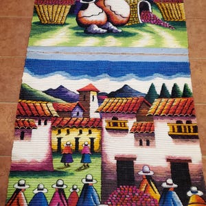 以下が含まれることがあります： カラフルな家、人物、紫色の農産物でいっぱいのバスケットがある村のシーンを描いた織りのタペストリー。このアートワークは、青い空、山、赤、青、黄色の鮮やかなカラーパレットを特徴としています。