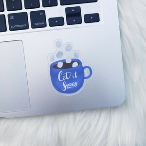 May include: A blue mug with white marshmallows and steam coming out of it. The mug has the text "Let it Snow" written on it.