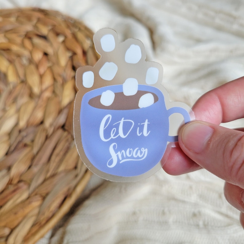 May include: A clear sticker with a blue mug filled with hot chocolate and marshmallows. The mug has the text "Let it Snow" written on it.