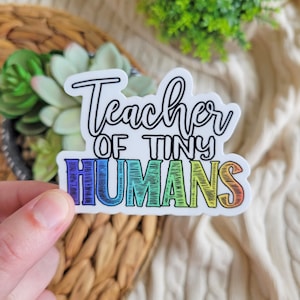 Puede incluir: Una pegatina de colores arcoíris con el texto "Teacher of Tiny Humans" en letra negra.