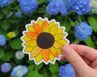 May include: A colorful sunflower sticker with yellow, orange, and brown details. The sticker has a white border and is held in front of a background of blue flowers and green leaves.