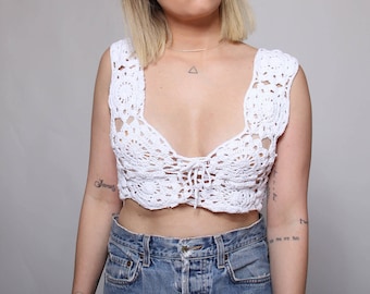 May include: White crochet crop top with a tie detail in the front. The top is made of a delicate, open-weave fabric and has a relaxed, bohemian style.